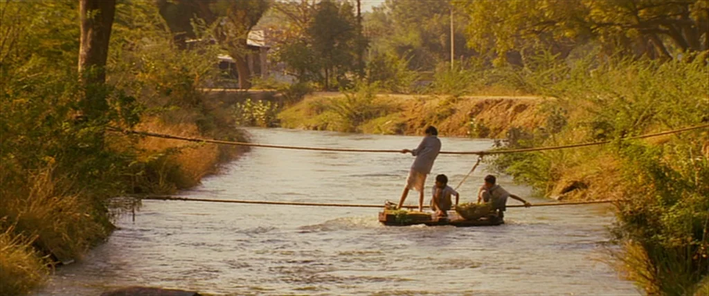 The Darjeeling Limited (2007) movie still: wide — Three boys navigate a simple wooden raft across a narrow, sunlit rural river. Dense foliage and…