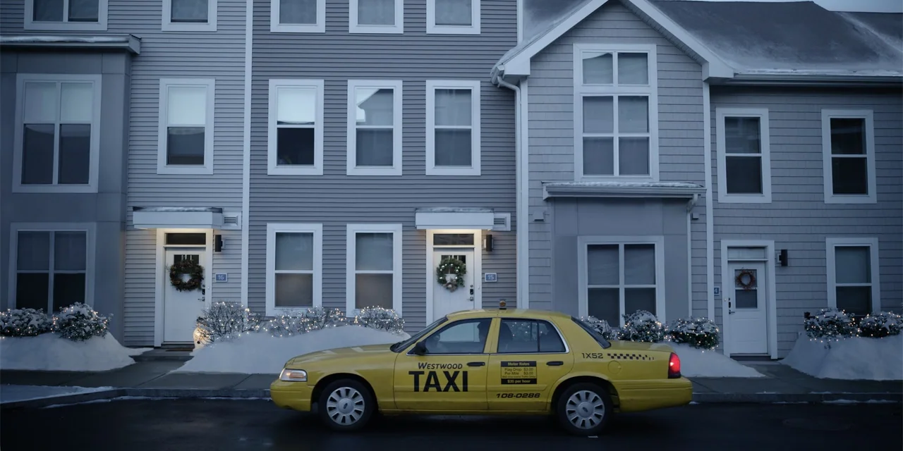 About Fate (2022) movie still: wide — A row of modern gray-sided suburban townhouses sits behind a snow-dusted front garden. A yellow taxi is parked…