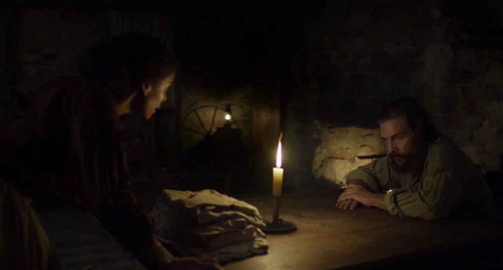 Free State of Jones (2016) movie still: medium — A dark, dimly lit rustic interior with stone walls and wooden furniture. A man and a woman sit at a…