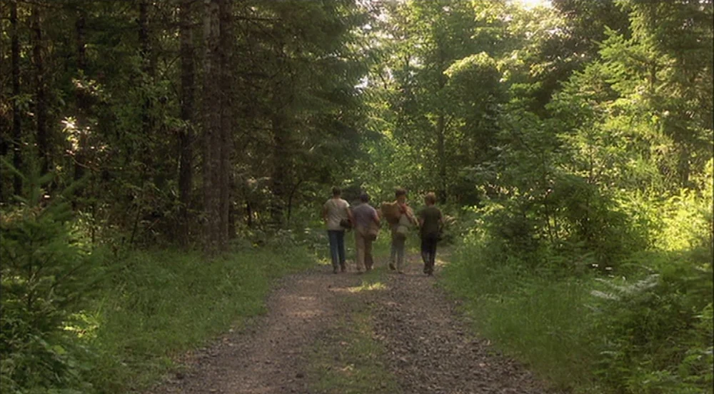 Stand By Me (1986) movie still: wide — A gravel trail cuts through a dense forest with tall pine and deciduous trees. Four boys walk away from the…