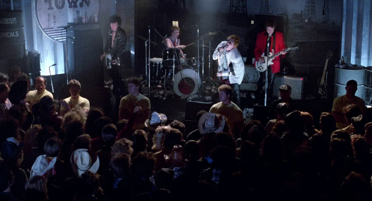 Sid and Nancy (1986) movie still: wide — A dimly lit indoor concert venue features a band performing on a small stage. A large, dense audience…