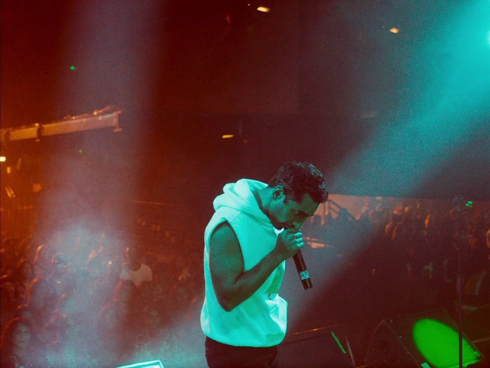Mogul Mowgli (2020) movie still: medium — A dimly lit concert stage filled with haze and saturated colored light beams. A man in a white sleeveless…