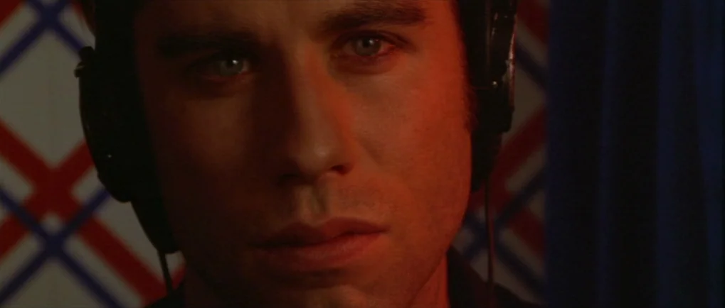 Blow Out (1981) movie still: closeup — A dark, cramped space with a patterned fabric wall behind the subject. A man wears large headphones while…