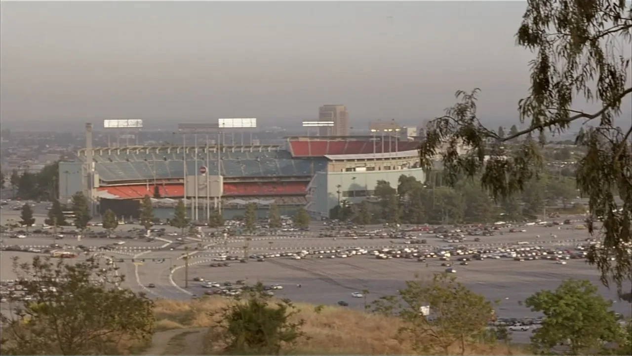 Ajuste de Cuentas (2016) movie still: establishing — An expansive outdoor view depicts a large sports stadium surrounded by a parking lot. Many cars…