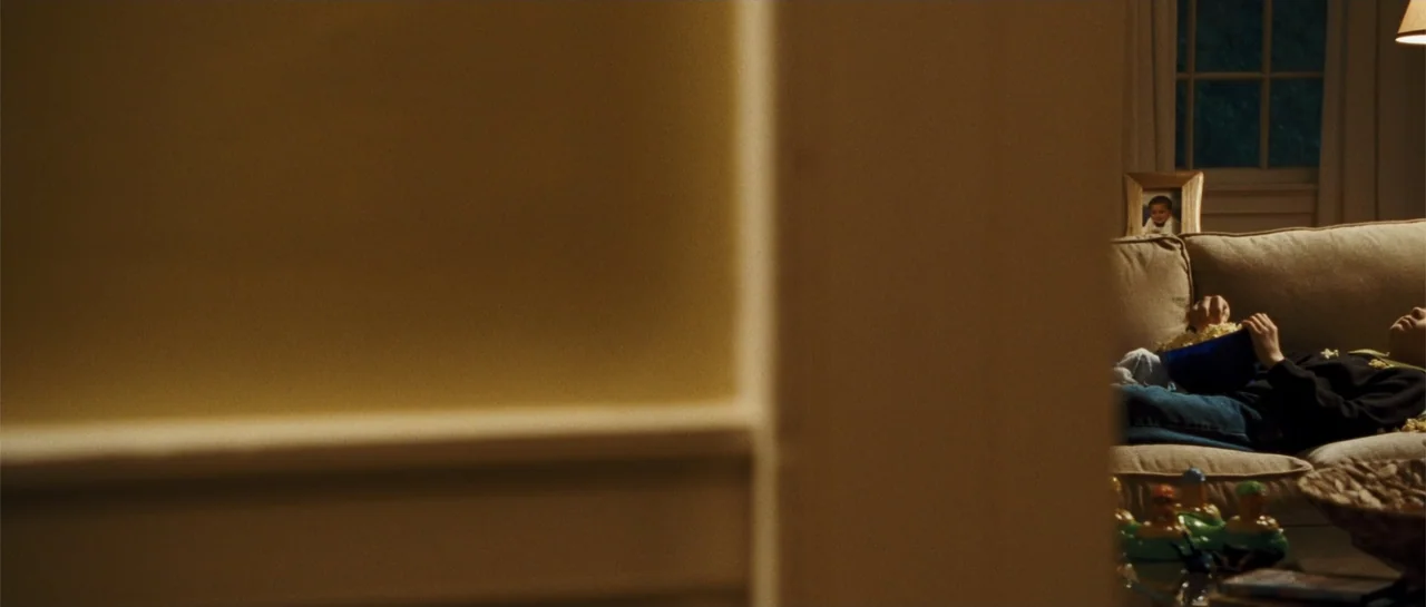 The New Daughter (2009) movie still: medium — A dark, partially obscured living room features a beige couch with two people sitting. They are eating…
