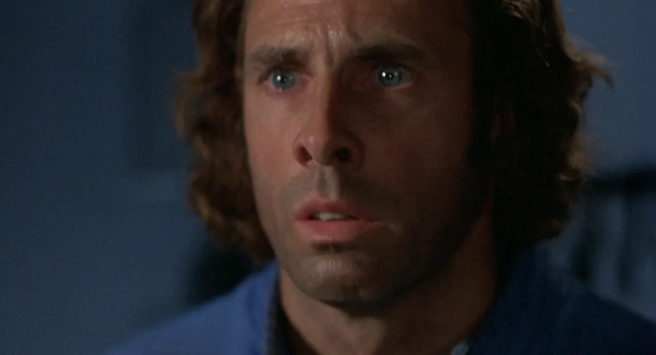 Silent Running (1972) movie still: closeup — A dimly lit indoor space with a plain dark wall backdrop. A man stares forward with wide eyes and a…