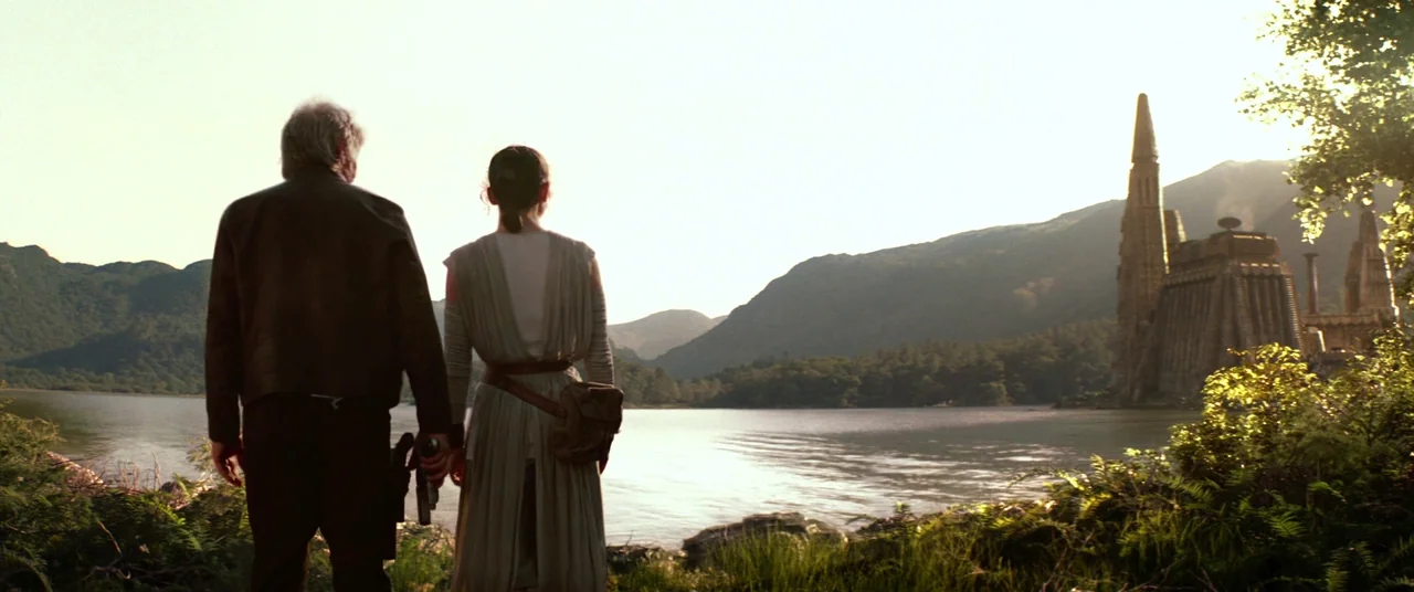 Star Wars: The Force Awakens (2015) movie still: wide — A man and a young woman stand on a grassy lakeshore looking toward a building. The building…