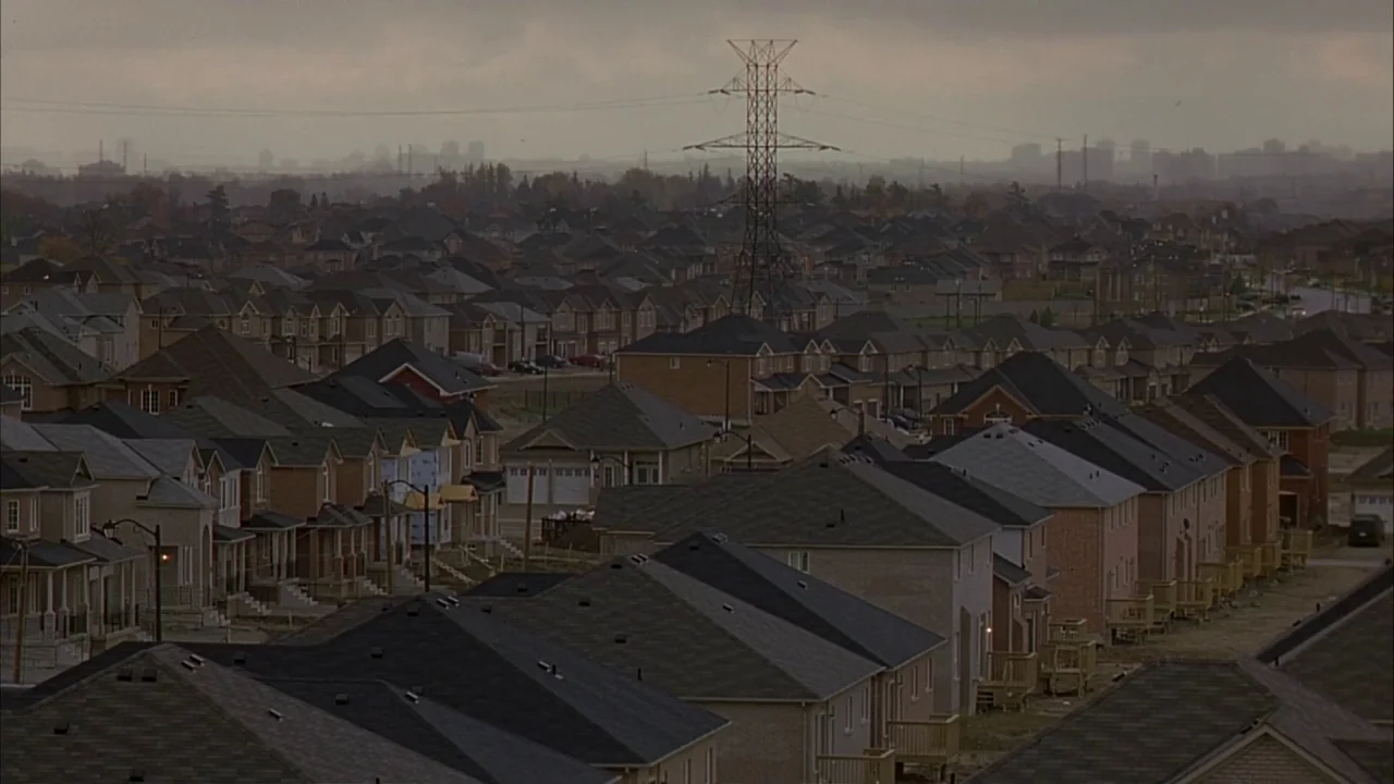 Ginger Snaps (2000) movie still: establishing — A vast suburban residential development fills the frame under a heavy, overcast gray sky. Rows of…