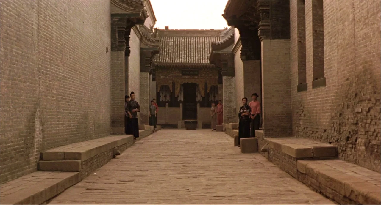 Raise The Red Lantern movie still: wide — A stone-paved courtyard is flanked by long grey brick walls and traditional ornate overhangs. Several…