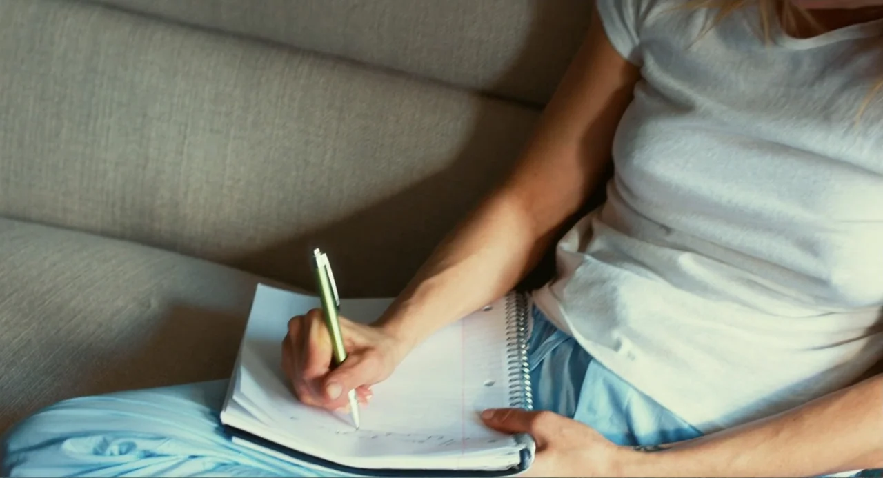 An Imperfect Murder (2017) movie still: medium — A person sits on a textured fabric sofa while holding a notebook. The subject uses a pen to write…