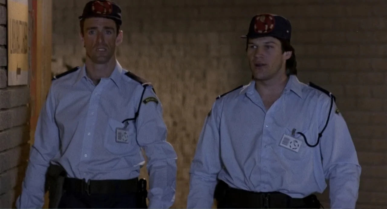 American Ninja 3: Blood Hunt (1989) movie still: medium — Two men walk through a generic commercial building corridor with plain brick walls. The…