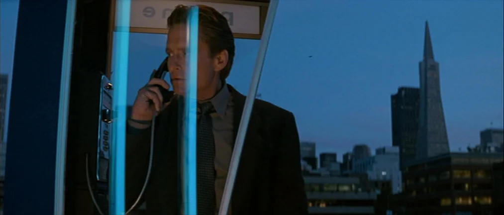 Basic Instinct (1992) movie still: medium — A man stands inside a glass phone booth overlooking a city skyline at dusk. The Transamerica Pyramid is…