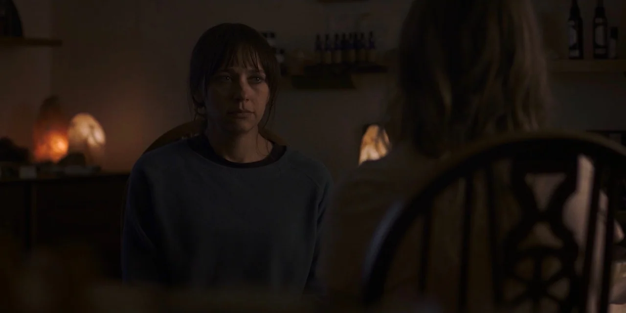 The Sound of Silence movie still: medium — A dimly lit room contains two people sitting at a table at night. One woman faces the camera directly…