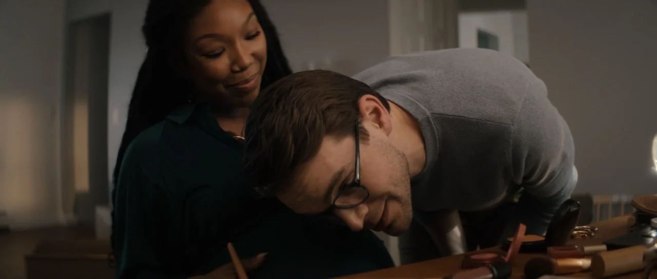 The Front Room (2024) movie still: medium — An interior bedroom space with cosmetics scattered on a wooden table surface. A man with glasses leans…