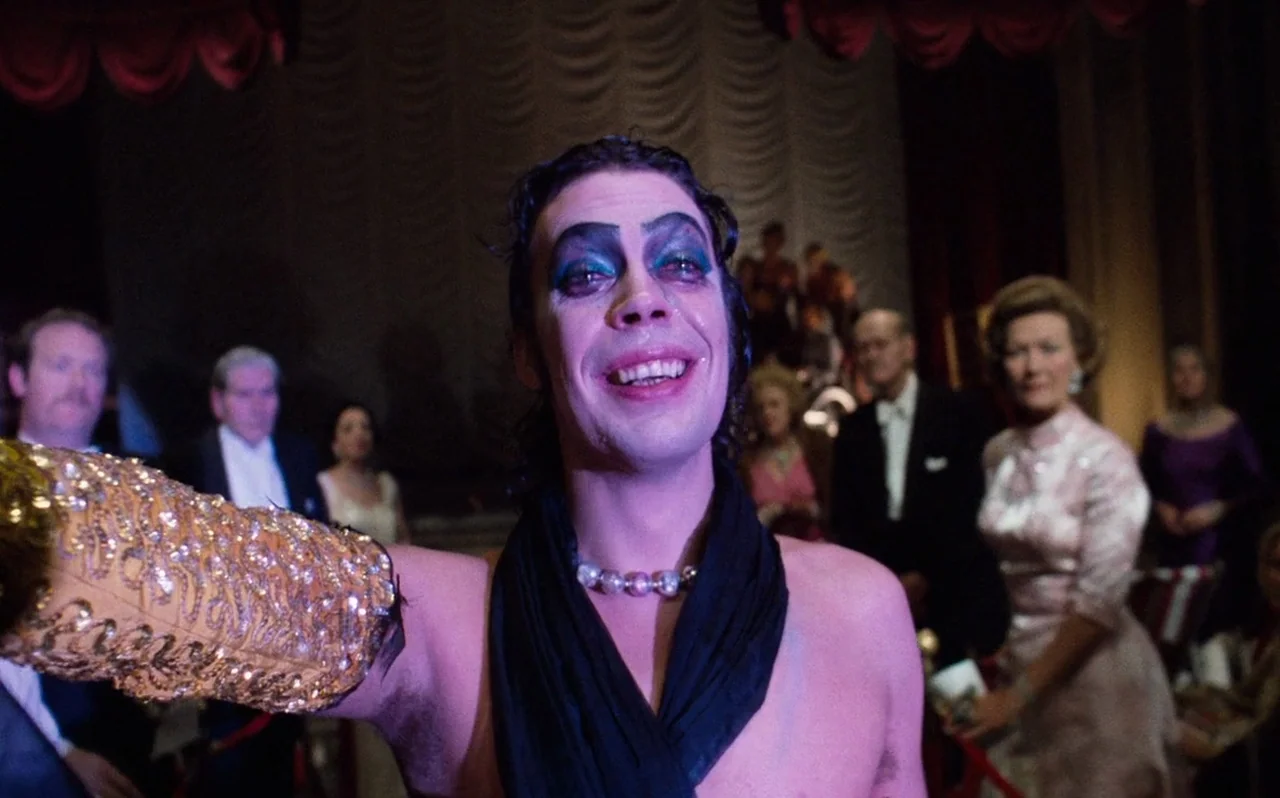 The Rocky Horror Picture Show (1975) movie still: medium — A dimly lit ballroom background with guests in formal attire standing behind the…