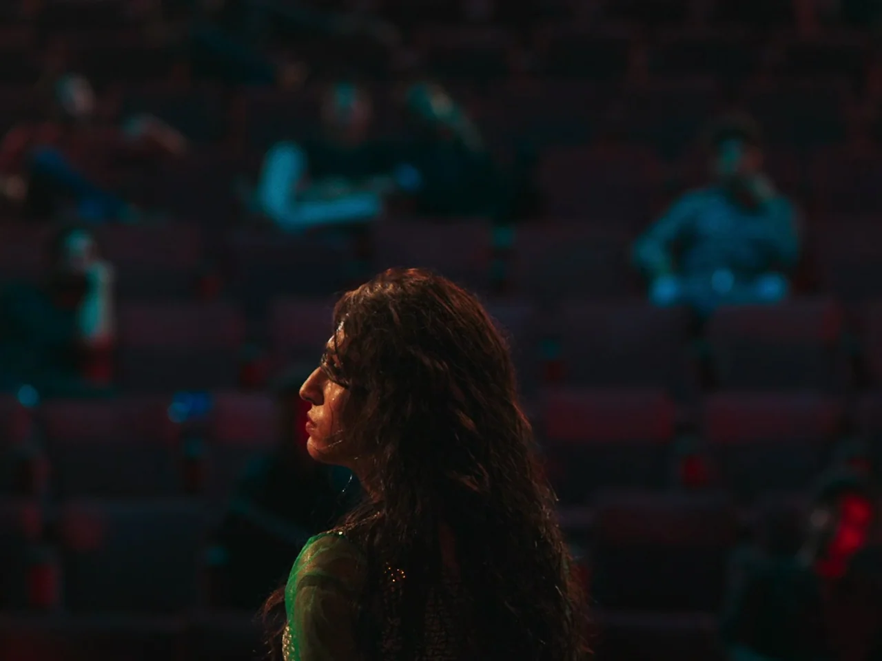 Joyland (2022) movie still: medium — A dimly lit movie theater with rows of dark, soft-backed seats. A young woman sits in the foreground, turned to…