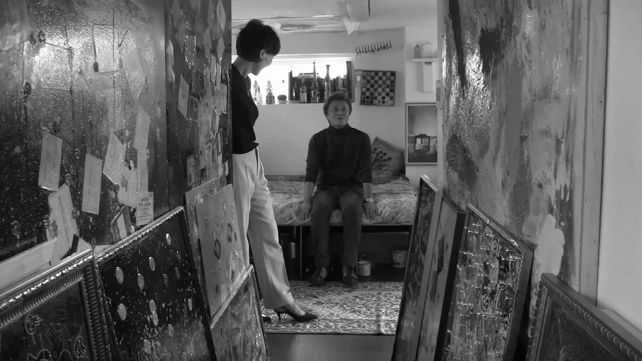 Walk Up (2022) movie still: fullbody — A cluttered apartment space features walls adorned with numerous framed artworks and textured boards. A woman…