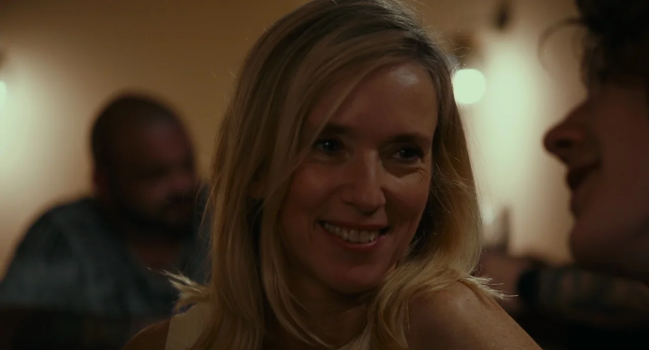 Last Summer (2023) movie still: closeup — A dimly lit indoor space with blurred figures in the background. A blonde woman smiles while engaged in…