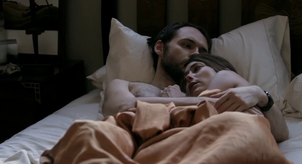 Sound Of My Voice (2011) movie still: medium — A man and woman lie together in a bed with white pillows. The pair is covered by a rust-colored…