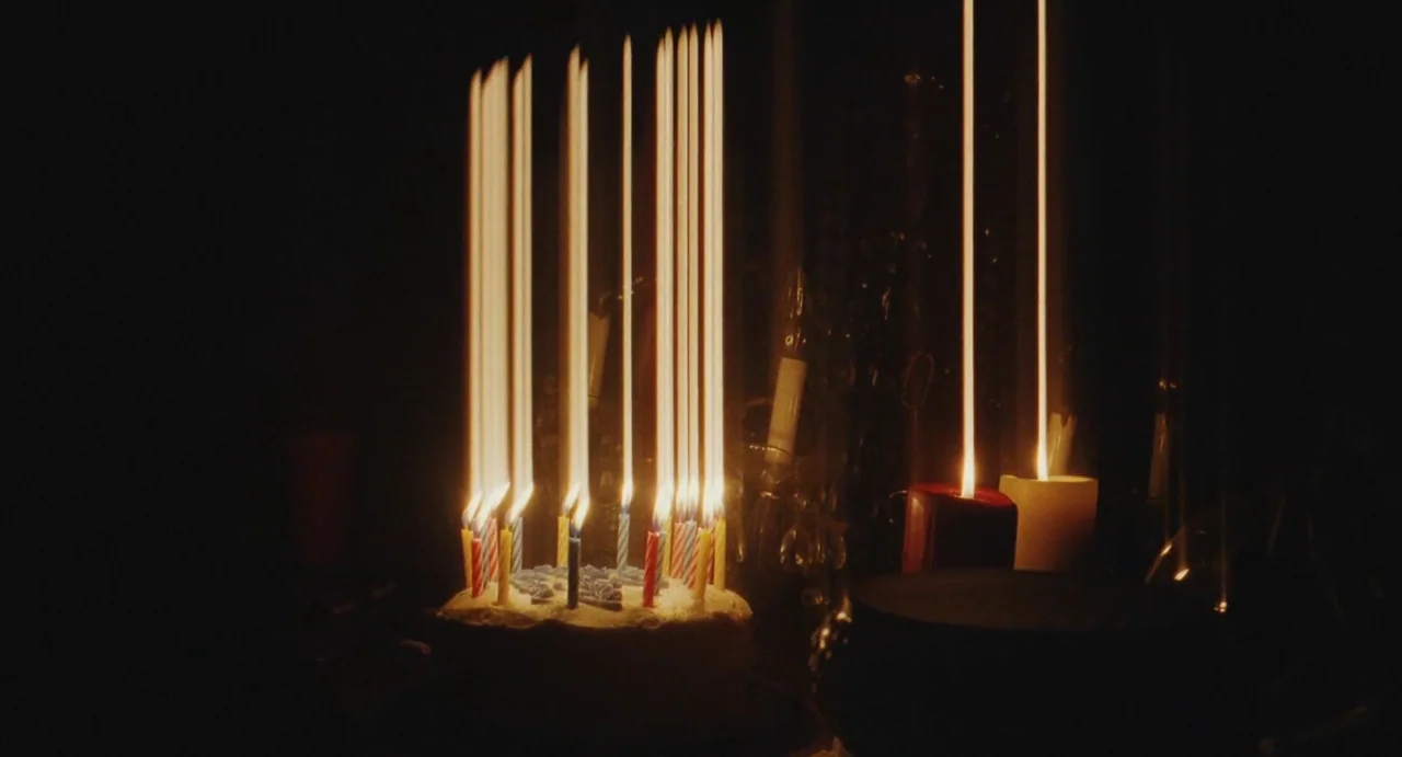 The Giant (2019) movie still: closeup — A dark, dimly lit space features a birthday cake topped with many lit candles. Long, vertical light…
