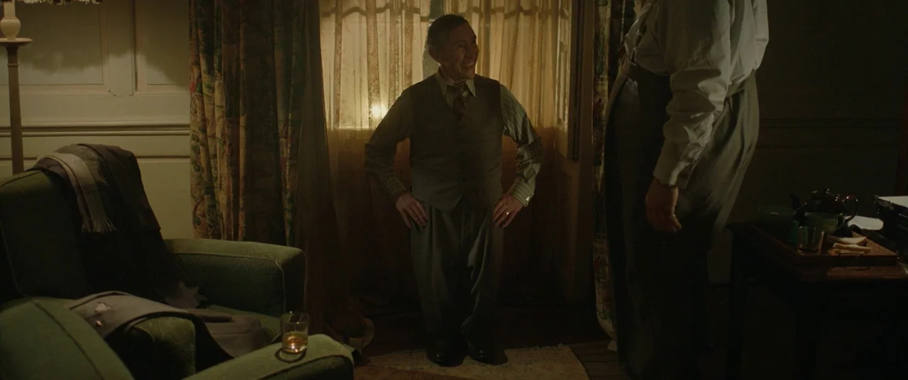 Domino (2005) movie still: fullbody — A dimly lit room features patterned curtains, a green armchair, and a side table. A short older man smiles at…