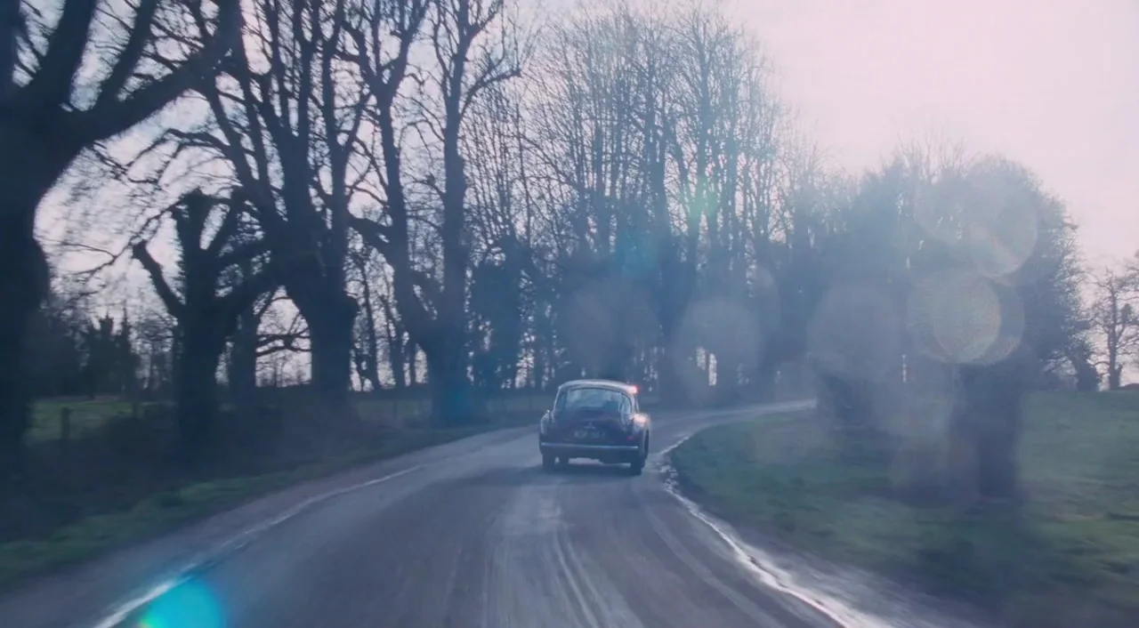 Phantom Thread (2017) movie still: wide — A rural road winds through an avenue of bare deciduous trees under overcast sky. A dark vintage car drives…
