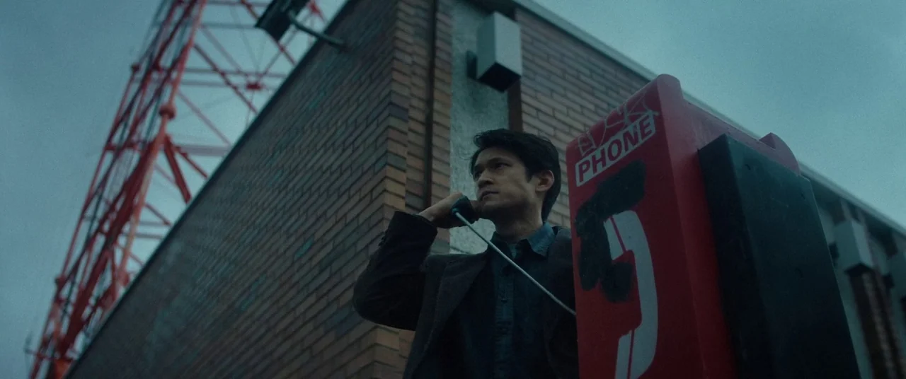 Broadcast Signal Intrusion (2021) movie still: medium — A brick wall and red telephone stand under a tall telecommunications transmission tower. The…