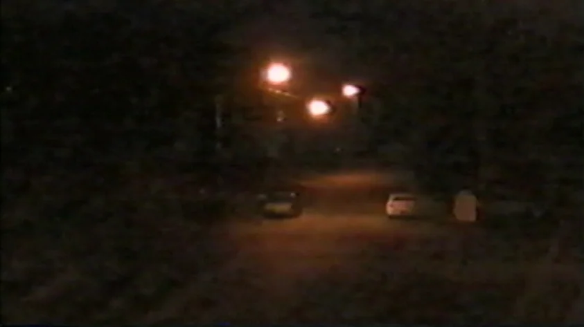 Trash Humpers (2009) movie still: establishing — A dark, blurry outdoor scene features a road illuminated by bright streetlamp light. Two small,…