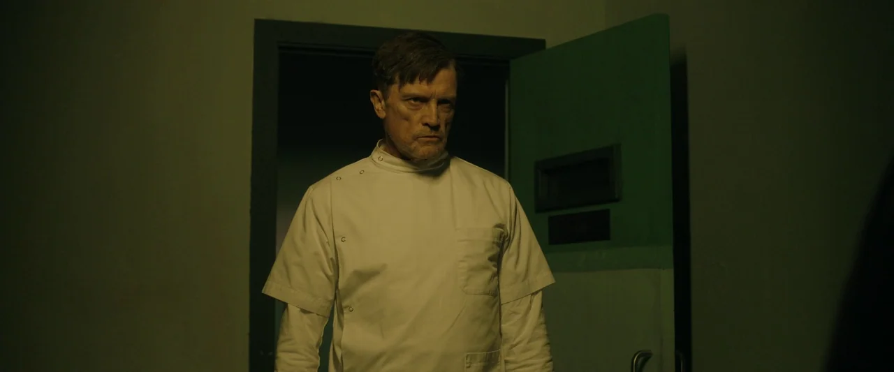 Oddity (2024) movie still: medium — A dimly lit industrial hallway features a green door with a small observation window. A man in a medical uniform…