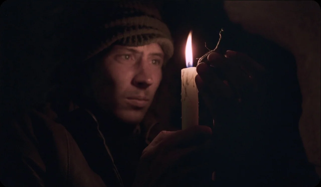 La Chimera (2023) movie still: closeup — A man stands in a dark space, his face partially illuminated by a candle. He watches the candle flame…