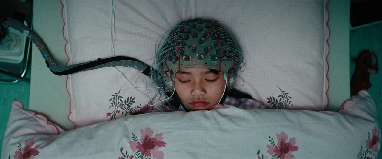 Cuckoo (2024) movie still: closeup — A girl lies in a hospital bed with a white pillow featuring floral patterns. She wears a medical EEG cap and is…