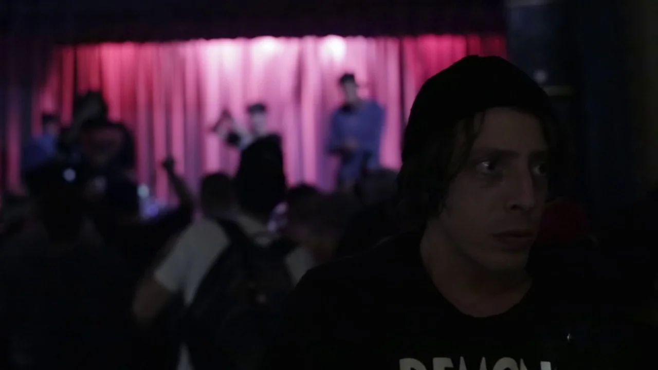 Buzzard (2014) movie still: closeup — A dark concert venue features a crowd and a brightly lit purple stage curtain. A young man in a beanie looks…