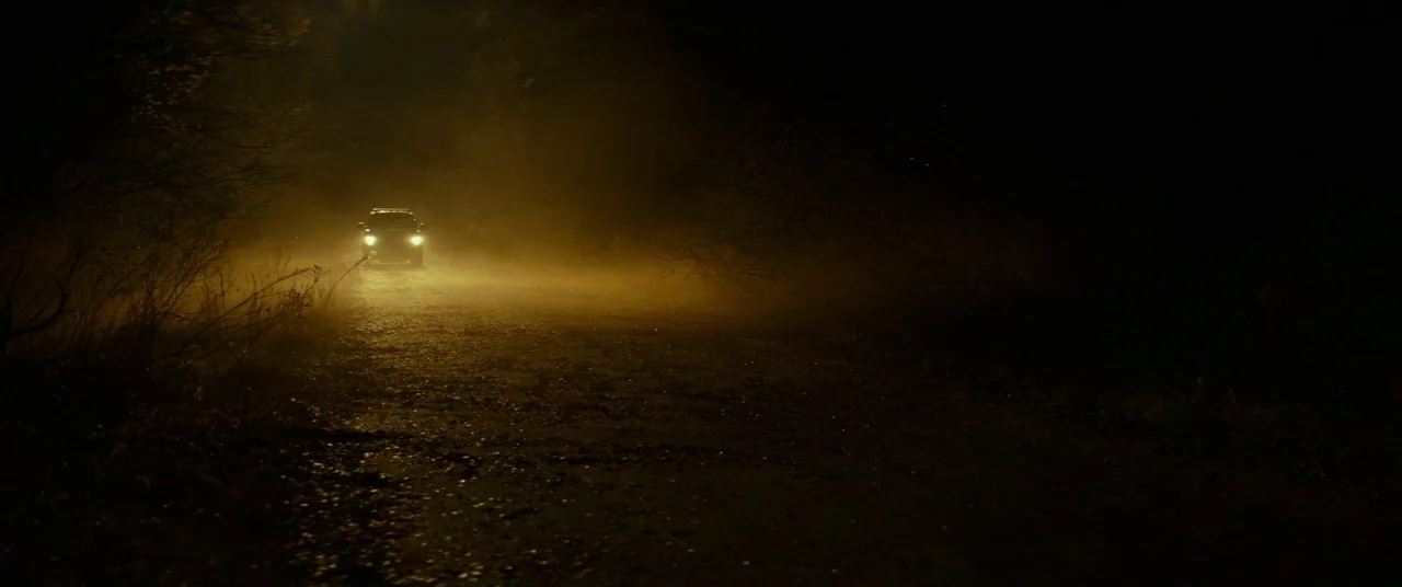 Halloween (2018) (2018) movie still: wide — A dark, unpaved rural road is surrounded by dense, shrouded vegetation on both sides. A lone vehicle…