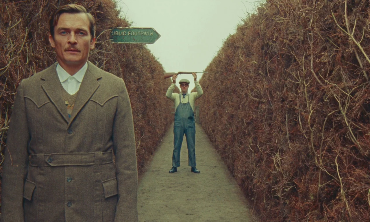 The Swan (2023) movie still: medium — A long narrow dirt path bordered by thick dry tall hedges. A man in the foreground looks forward while a man…