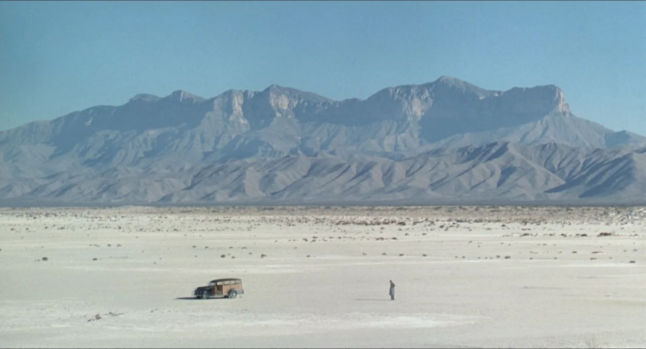 Lolita (1997) movie still: wide — A vast, flat, light-colored dry lake bed sits before an expansive mountain range. A lone man stands next to a…
