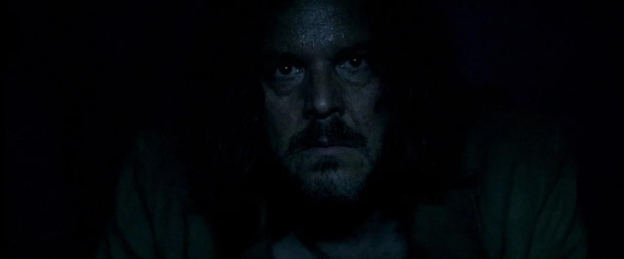 The Proposition (2005) movie still: closeup — A dark, dimly lit interior space with the subject centered against a black background. The subject is…
