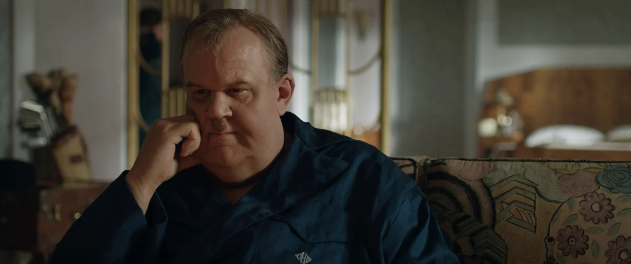 Stan & Ollie (2018) movie still: medium — A furnished hotel room features a patterned fabric sofa and decorative gold-framed room dividers. A man in…