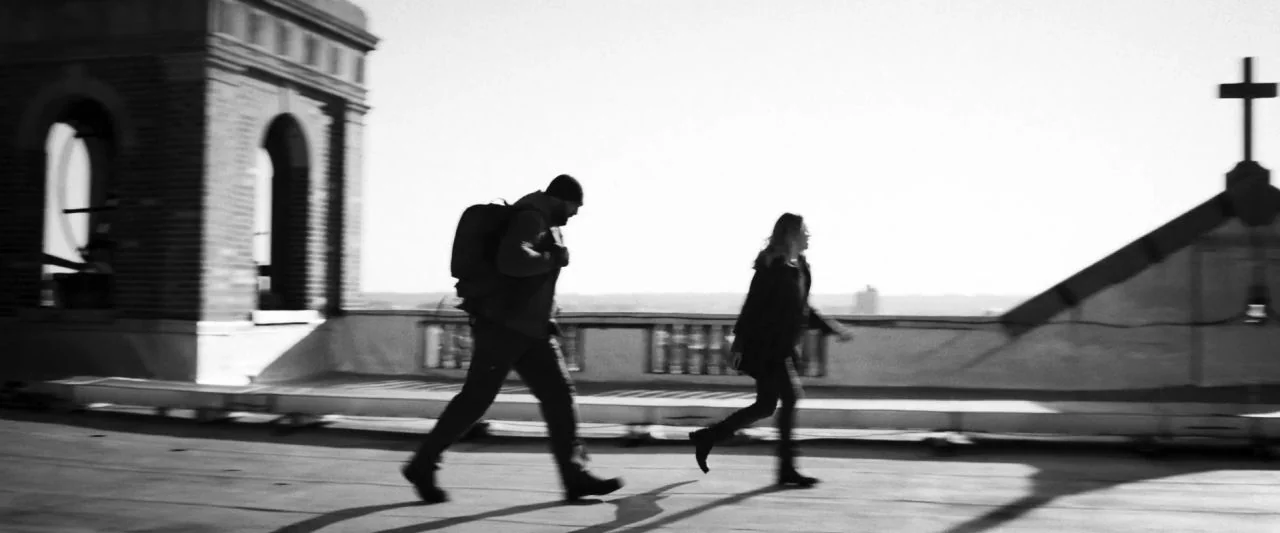 Bushwick (2017) movie still: wide — A flat rooftop with a brick chimney and a stone cross in the background. A man carrying a backpack and a woman…