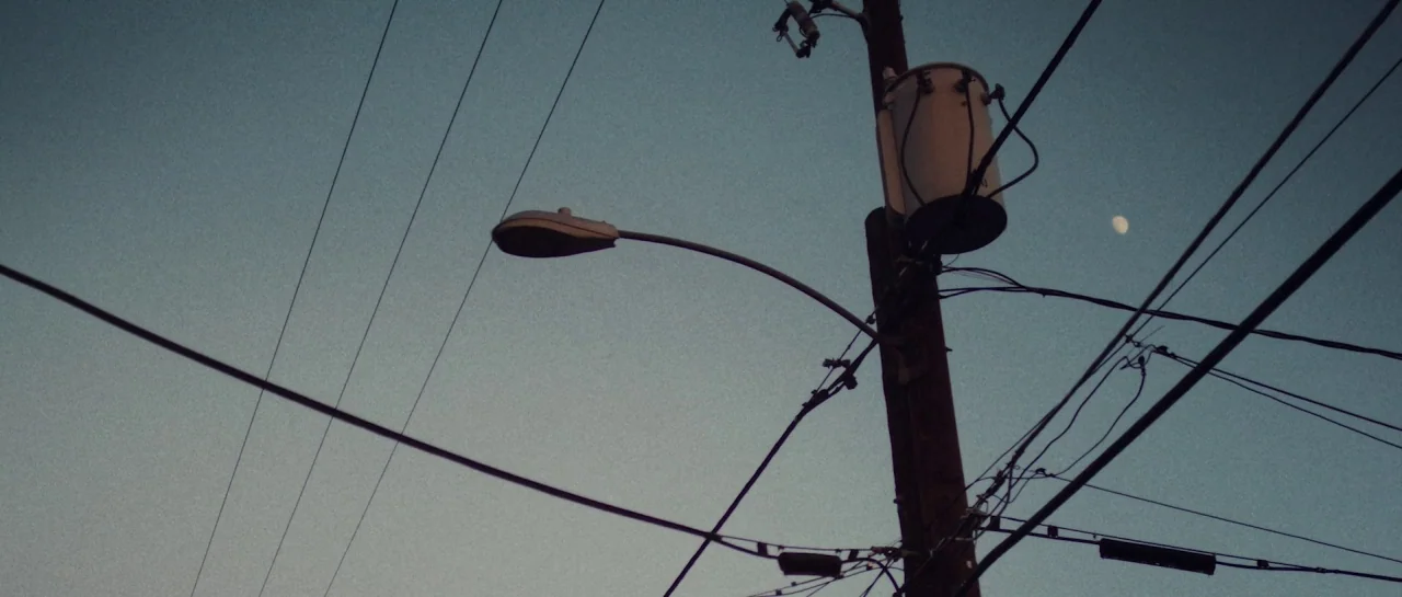 Starfish (2018) movie still: establishing — An urban utility pole stands against a clear, darkening blue twilight sky. Electrical wires and a street…