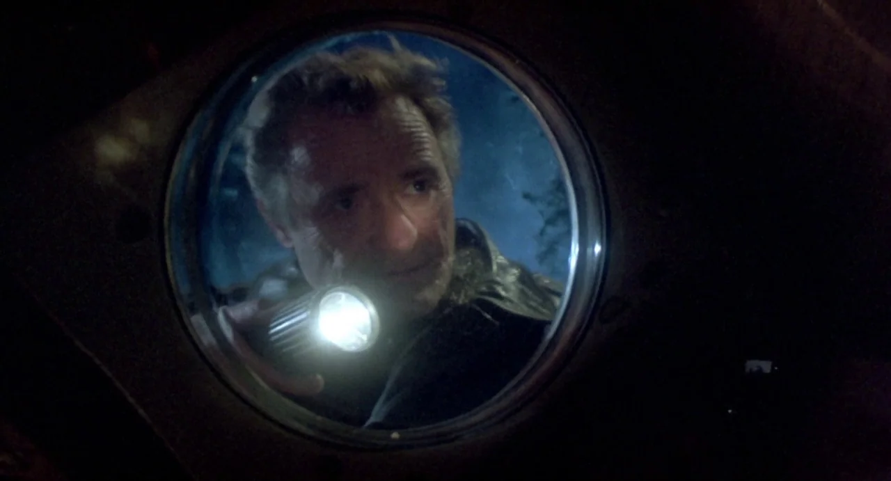 Explorers (1985) movie still: closeup — A circular window provides a view of a man peering through at night. The man holds a bright flashlight that…