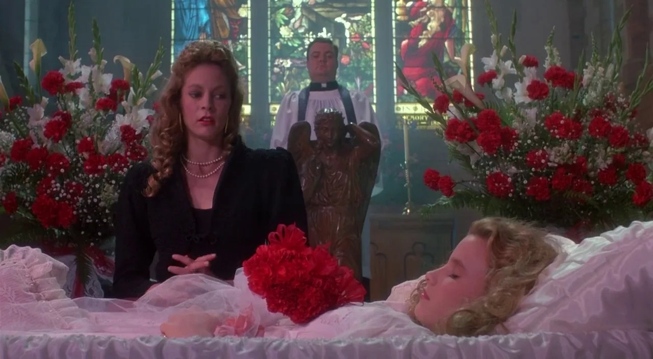 Heathers movie still: medium — A church chapel interior features stained glass windows and large red floral arrangements. A woman sits by a casket…