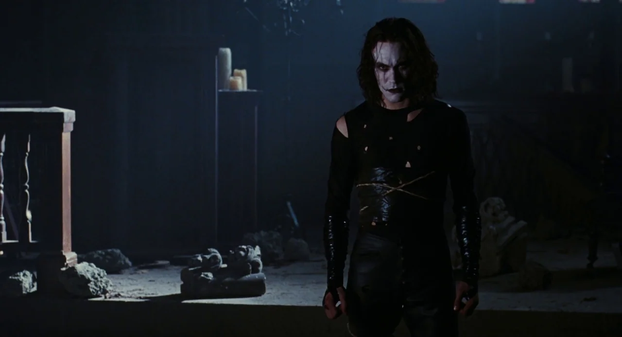 The Crow (1994) movie still: medium — A dimly lit, damaged stone interior space features scattered rubble and flickering candles. A man with…