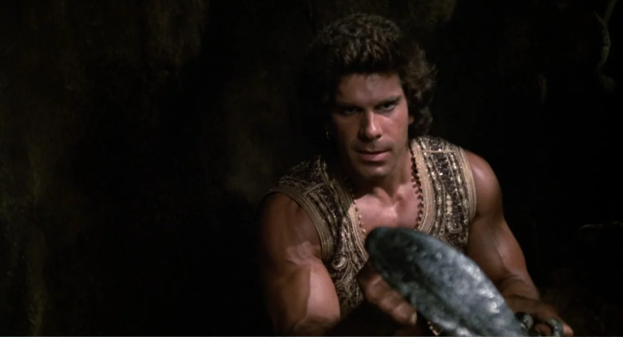 Hercules (1983) movie still: medium — A dark, rocky interior space serves as the backdrop for a muscular man. The man in an ornate vest holds an…