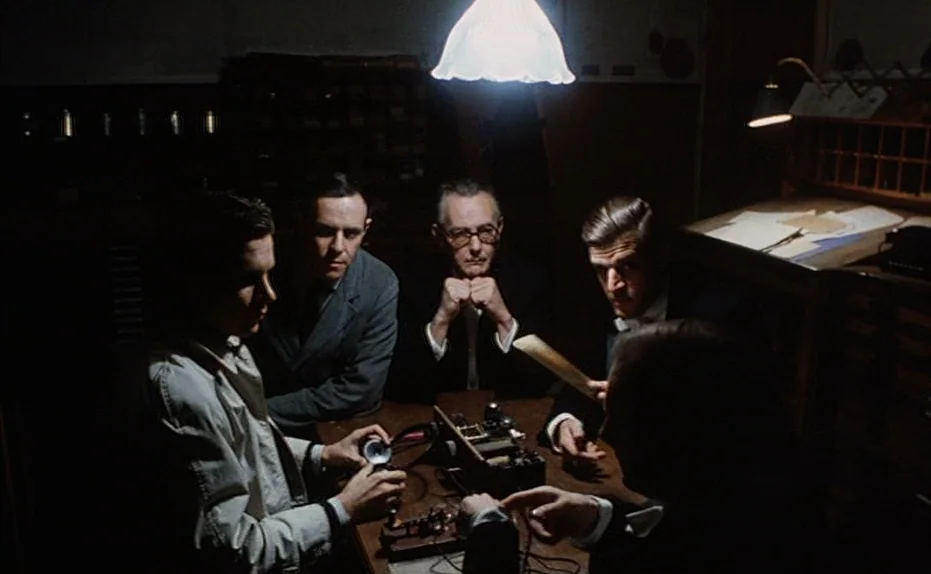 Soldier of Orange (1977) movie still: medium — Four men sit at a dark wooden table in a dimly lit, cluttered technical room. They focus on a small,…