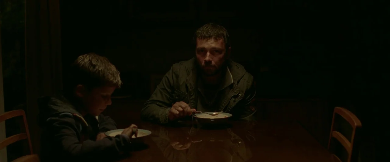 The End We Start From (2023) movie still: medium — A dark dining room features a polished wooden table with two people eating. The man stares…