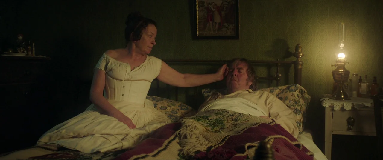 Mr. Turner (2014) movie still: medium — A dimly lit 19th-century bedroom features patterned green wallpaper and a brass bed frame. A woman in a…