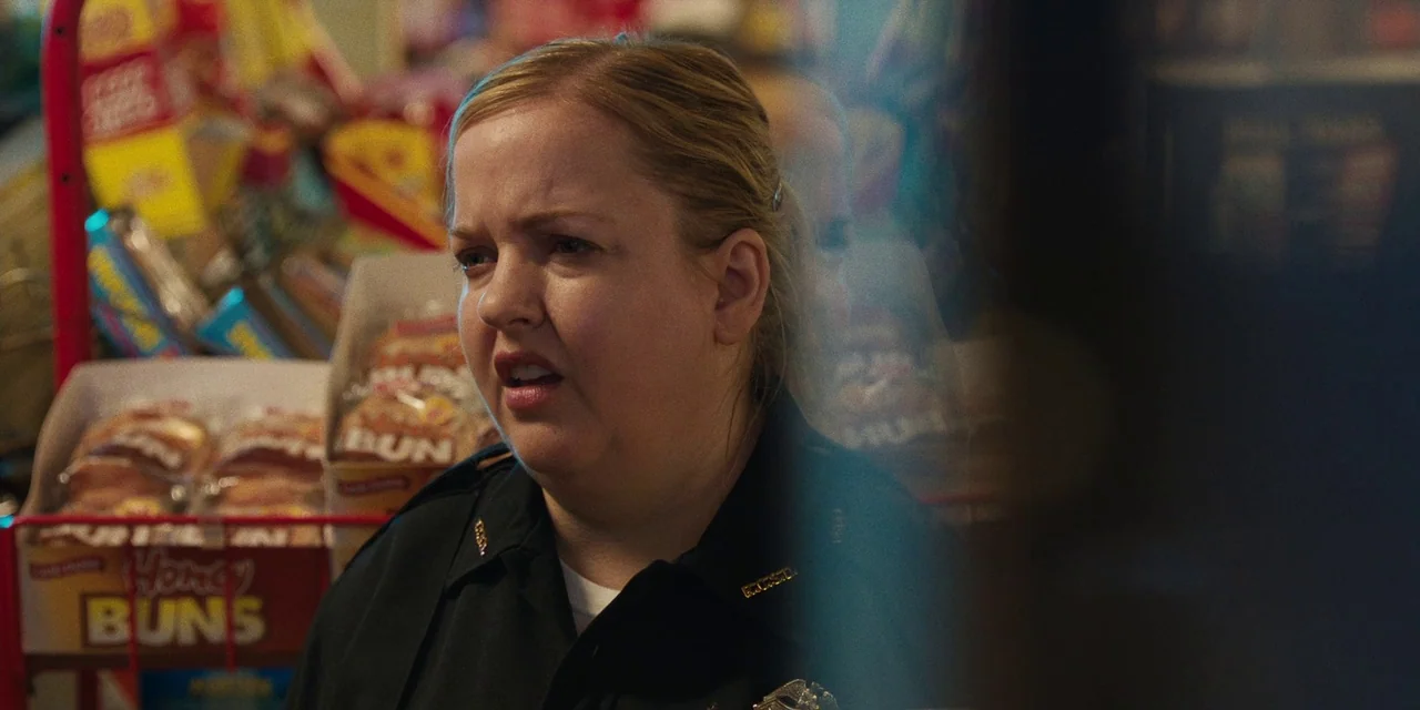 The Death of Dick Long (2019) movie still: closeup — A store aisle filled with boxes of baked goods stacked on a wire rack. A woman in a police…