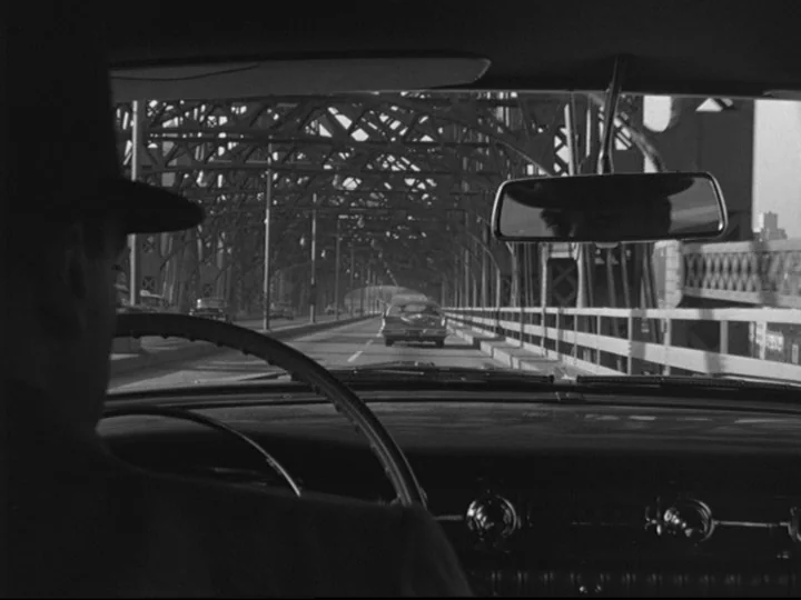Blast Of Silence (1961) movie still: A moody, noir-inspired shot from inside a car driving across a geometric metal truss bridge. The driver,…