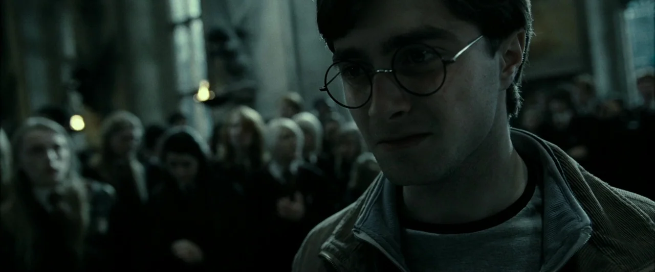 Harry Potter & The Deathly Hallows Part 2 (2011) movie still: closeup — A stone-walled interior hall with blurred figures standing in the…
