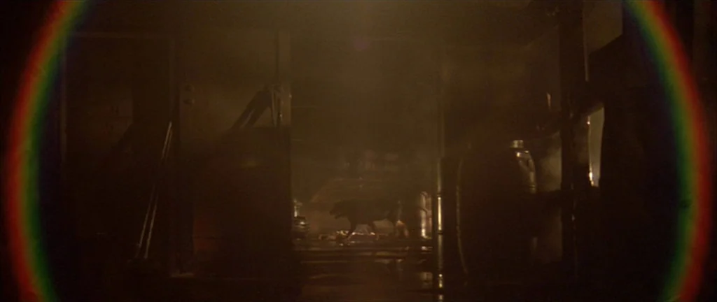 Alien 3 (1992) movie still: wide — A dark, industrial warehouse space contains stacked metal barrels and indistinct mechanical equipment. A dog…